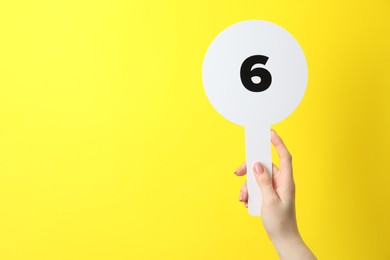 Photo of Woman holding auction paddle with number 6 on yellow background, closeup. Space for text