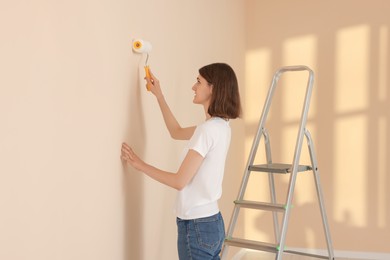 Photo of Young woman painting wall with roller near stepladder indoors. Room renovation
