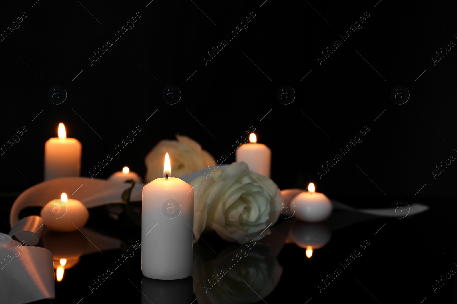 Photo of White roses and burning candles on black mirror surface in darkness, space for text. Funeral symbols