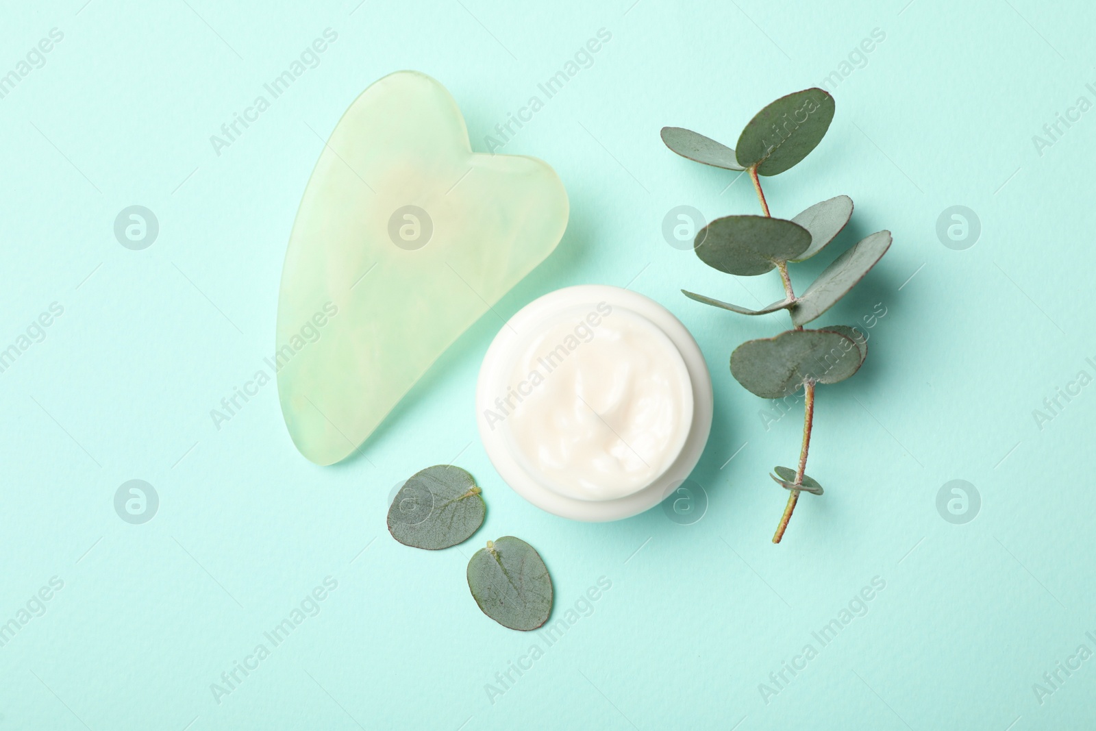 Photo of Jade gua sha tool, cream and eucalyptus branch on light blue background, flat lay