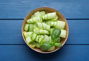 Slices of fresh ripe cucumber and basil in bowl on blue wooden table, top view