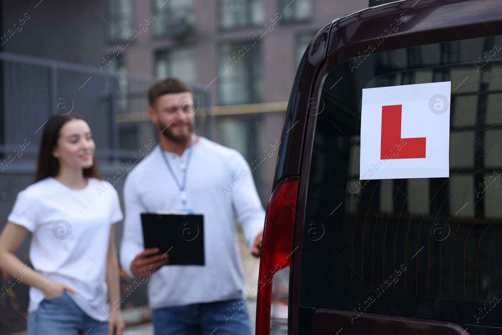 Photo of Learner driver and instructor with clipboard near car outdoors, selective focus on L-plate. Driving school