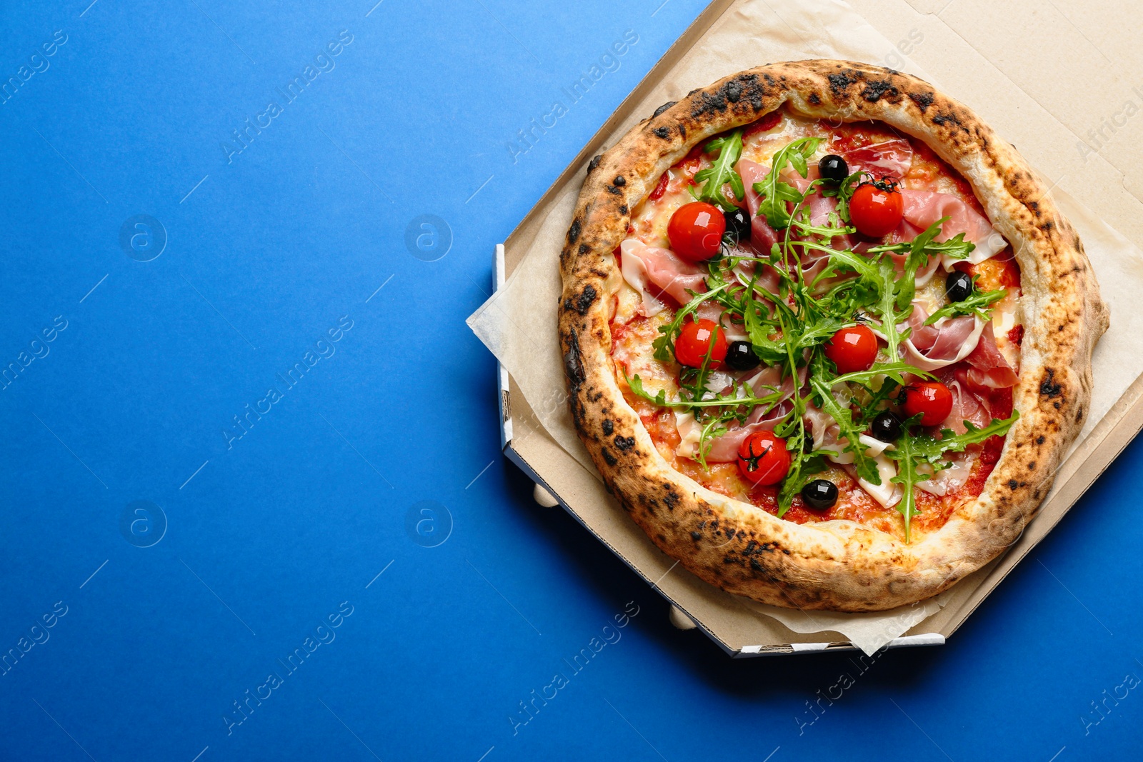 Photo of Tasty pizza with meat and arugula in cardboard box on blue background, top view. Space for text