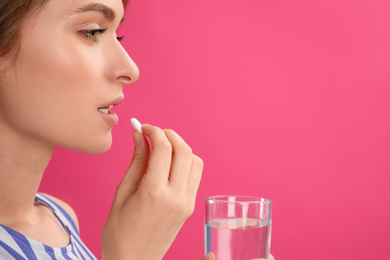 Young woman with glass of water taking vitamin pill on pink background. Space for text