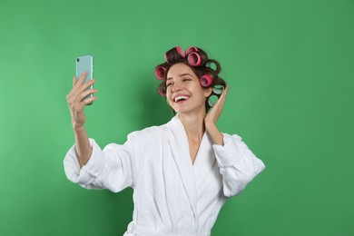 Photo of Happy young woman in bathrobe with hair curlers taking selfie on green background