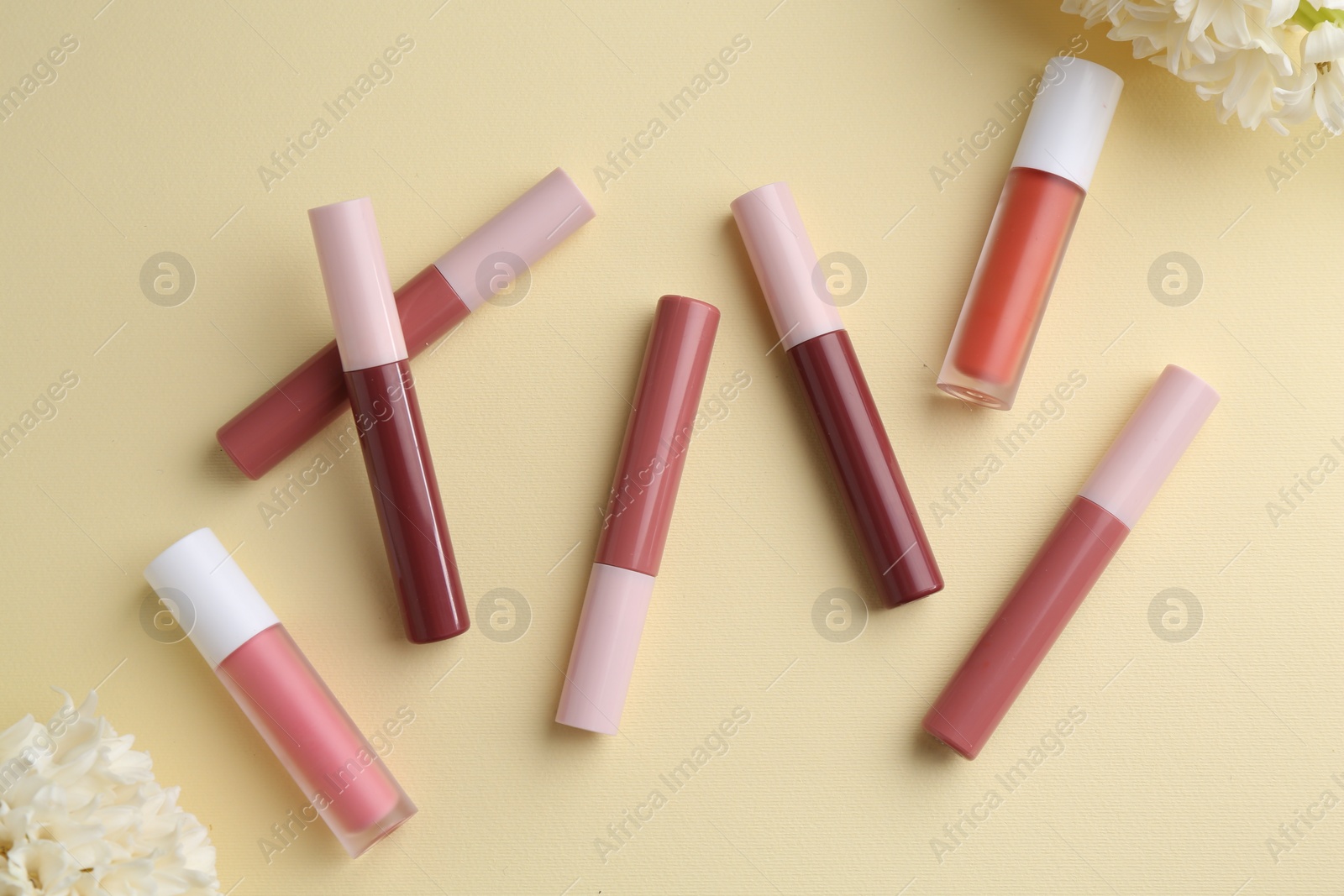 Photo of Different lip glosses and flowers on pale yellow background, flat lay