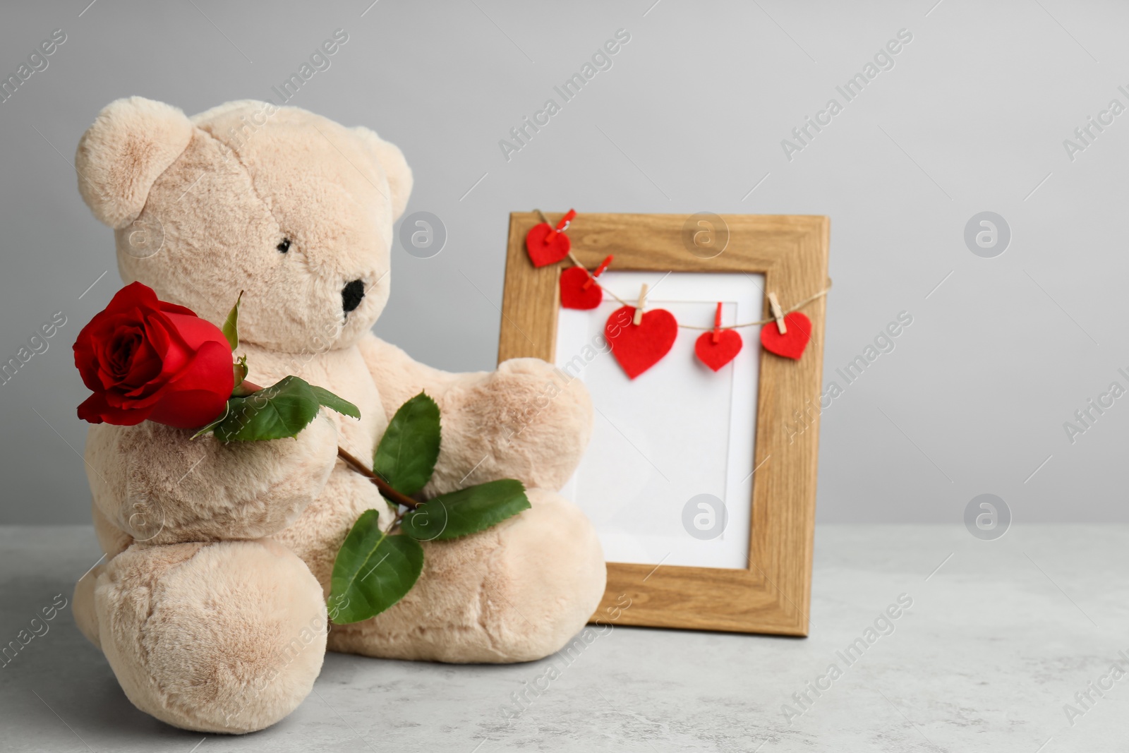 Photo of Cute teddy bear with red rose and frame on light grey stone table. Valentine's day celebration