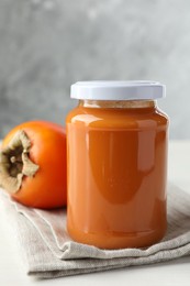 Photo of Delicious persimmon jam in glass jar and fresh fruit on white table
