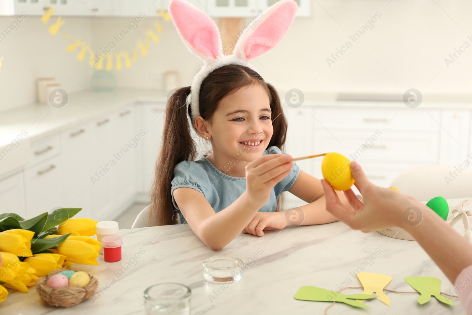 Photo of Happy little girl with bunny ears headband and her mother painting Easter egg in kitchen