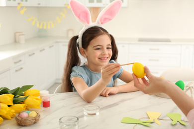 Happy little girl with bunny ears headband and her mother painting Easter egg in kitchen
