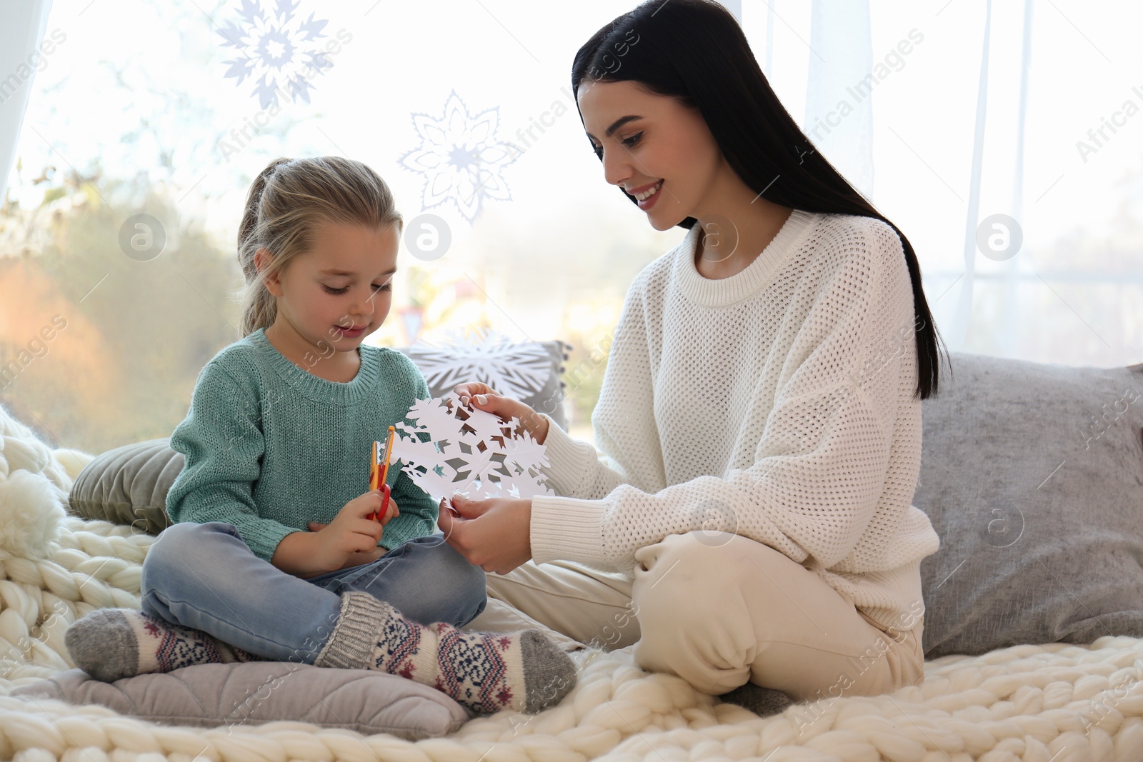 Photo of Mother and daughter making paper snowflake near window at home