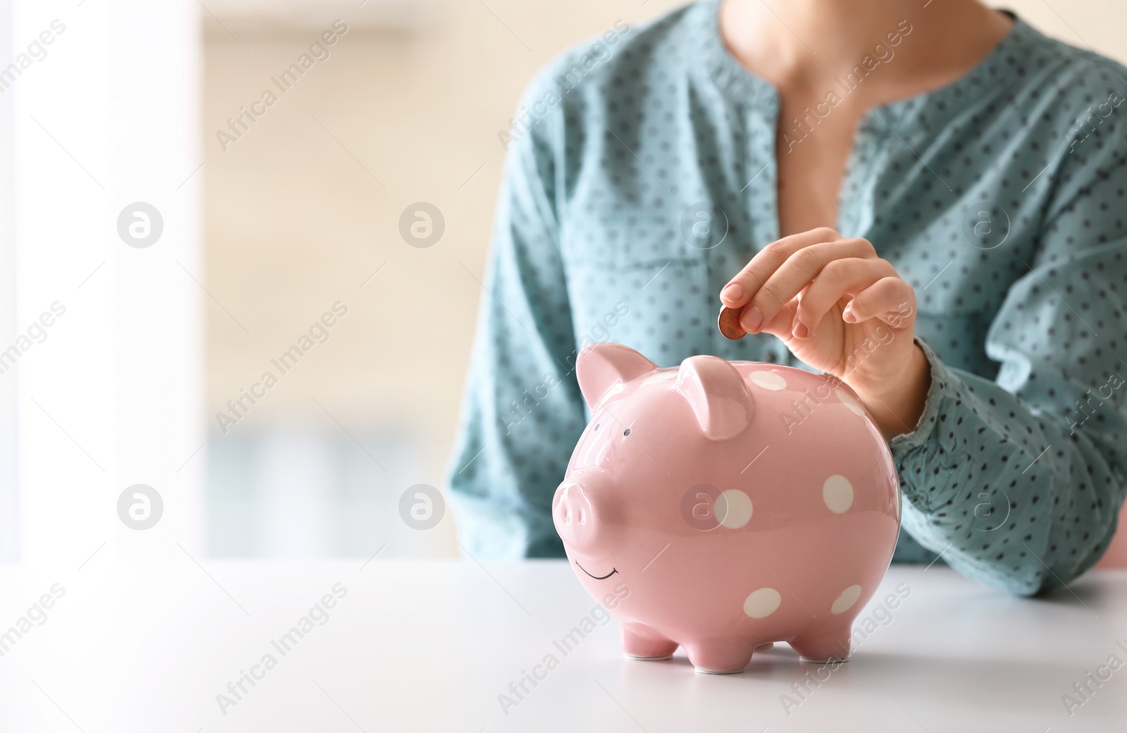 Photo of Woman putting coin into piggy bank at table indoors, closeup. Space for text