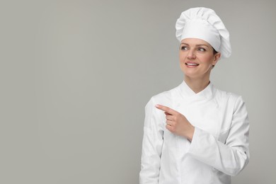 Photo of Happy woman chef in uniform pointing at something on grey background, space for text