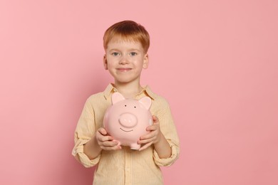Cute little boy with ceramic piggy bank on pale pink background