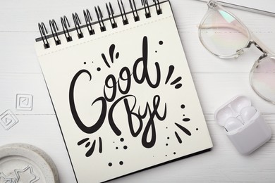 Image of Notebook with word Goodbye, eyeglasses and earphones on white wooden table, flat lay