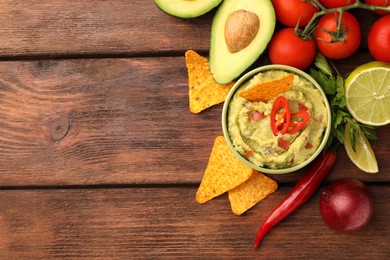 Photo of Bowl of delicious guacamole, nachos chips and ingredients on wooden table, flat lay. Space for text