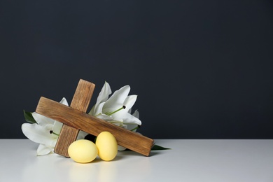 Photo of Wooden cross, Easter eggs and blossom lilies on table against color background, space for text