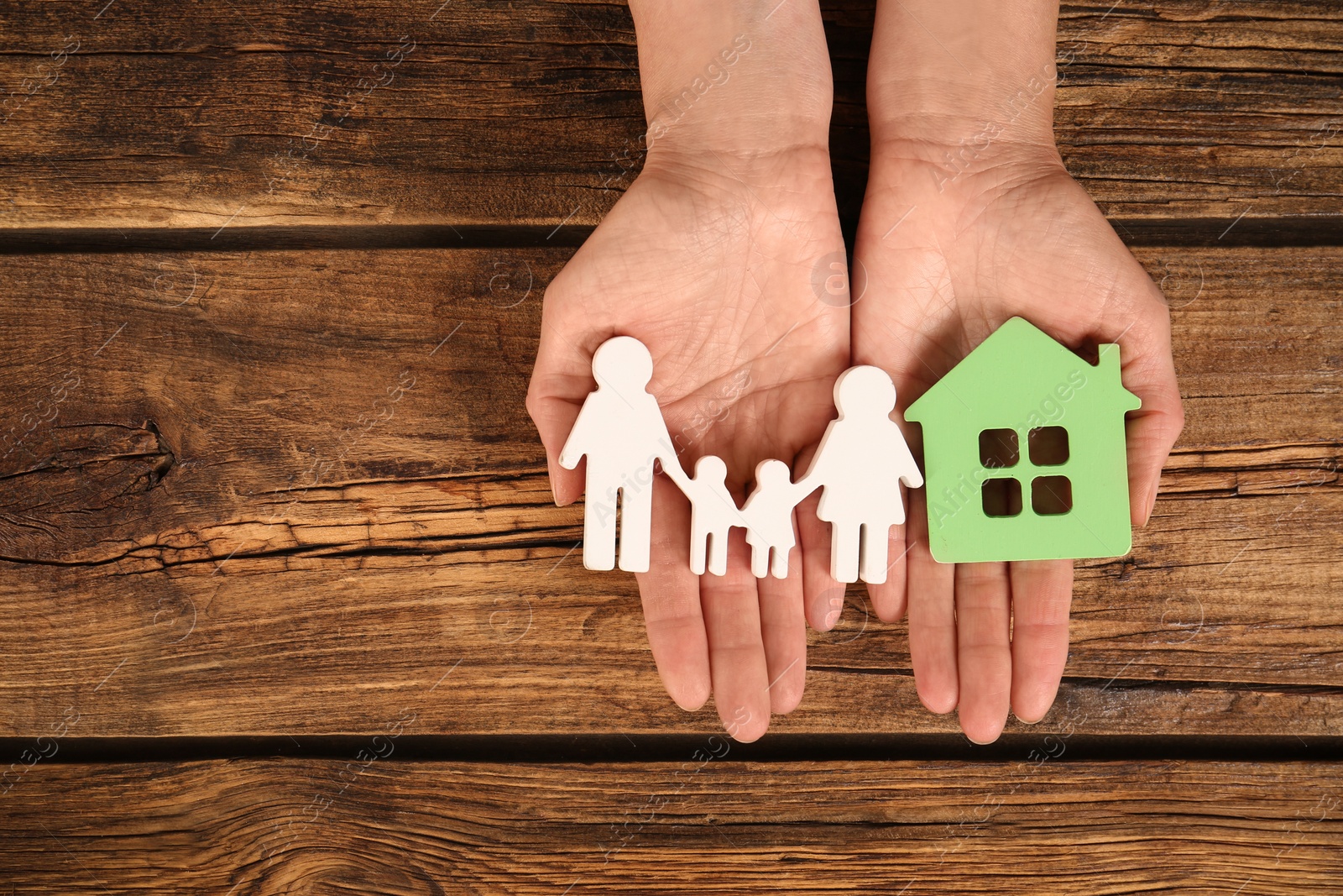 Photo of Woman holding figures of family and green house in hands on wooden background, top view. Space for text