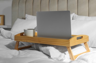 Wooden tray with modern laptop and cup of aromatic drink on bed indoors