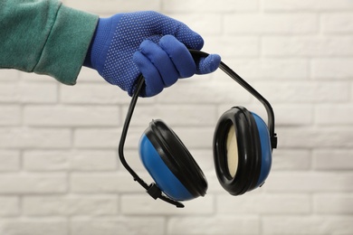 Worker holding safety headphones against white brick wall, closeup. Hearing protection device