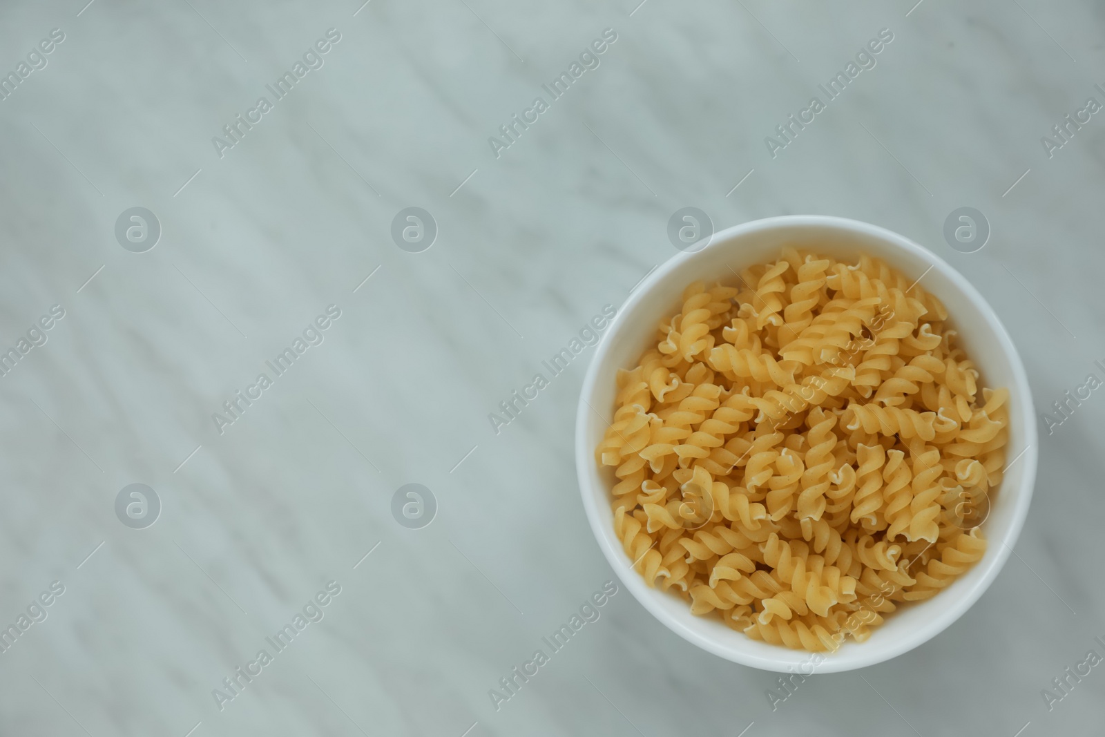 Photo of Bowl with uncooked fusilli pasta on white marble table, top view. Space for text