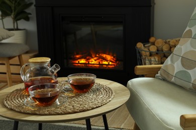 Teapot and cups of drink on coffee table near stylish fireplace in cosy living room. Interior design