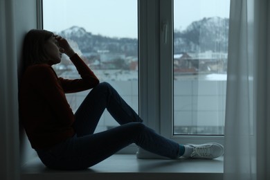 Photo of Sad young woman sitting on windowsill near window at home