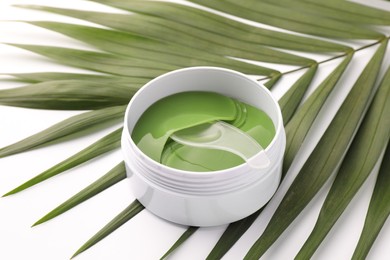 Photo of Jar of under eye patches with spoon and palm leaf on white background, closeup. Cosmetic product