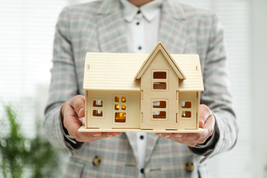 Real estate agent holding wooden house model with lights indoors, closeup