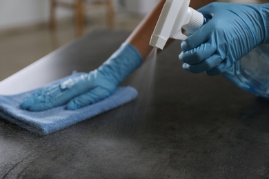 Photo of Woman in gloves cleaning grey stone table with rag and detergent indoors, closeup