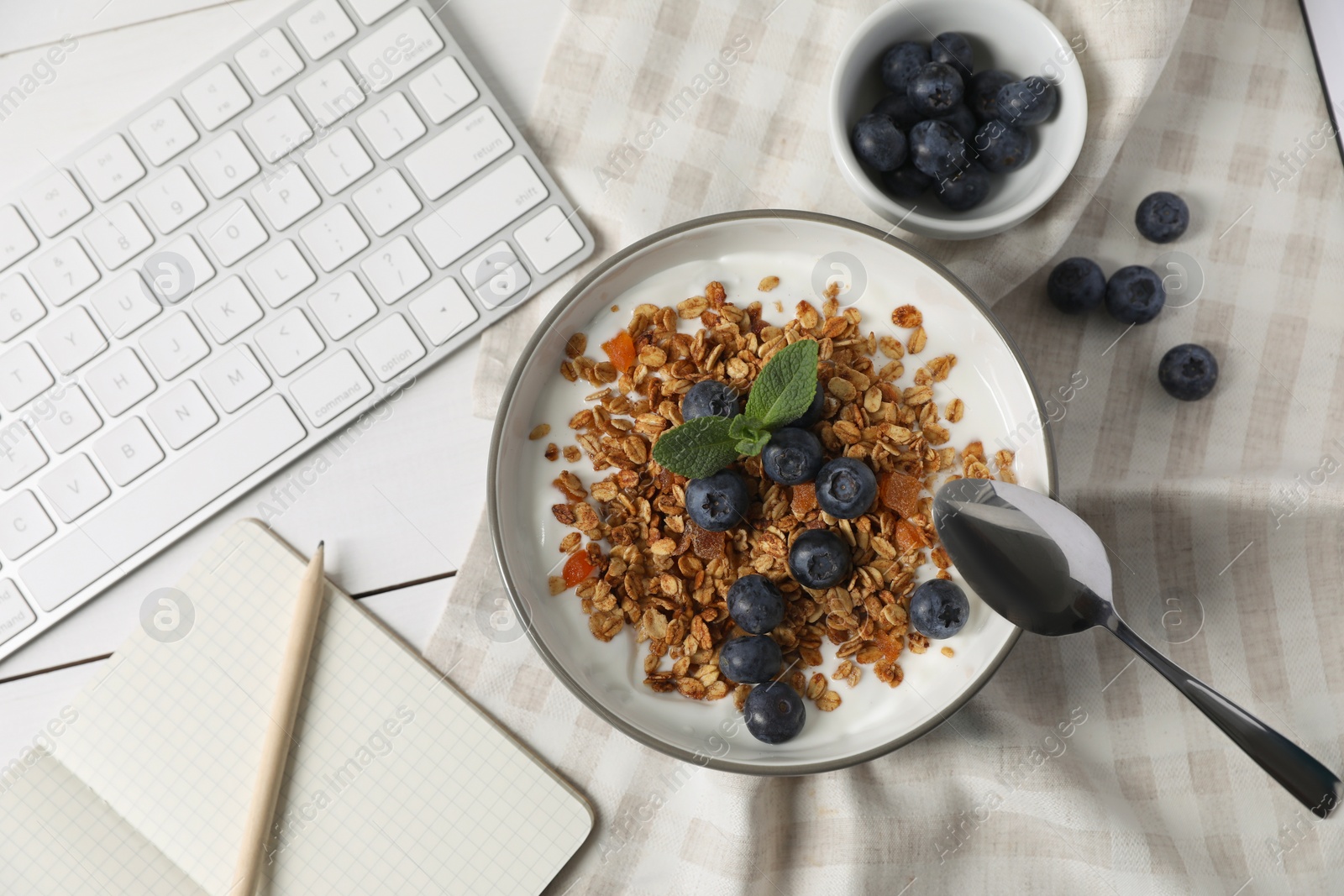 Photo of Delicious granola with blueberries in bowl, stationery and computer keyboard on white wooden table, flat lay