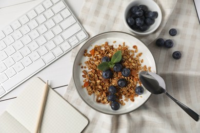 Delicious granola with blueberries in bowl, stationery and computer keyboard on white wooden table, flat lay