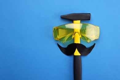 Photo of Man's face made of artificial mustache, safety glasses and hammer on blue background, top view. Space for text