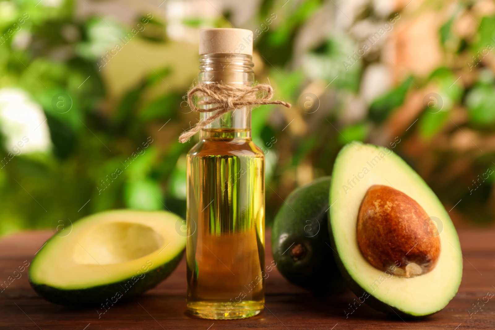 Photo of Glass bottle of cooking oil and fresh avocados on wooden table against blurred green background