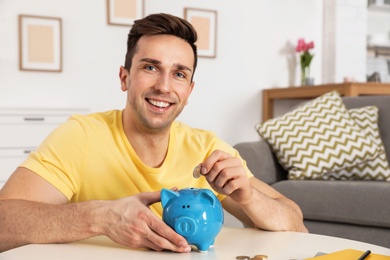 Photo of Happy man putting coin into piggy bank at table in living room. Saving money