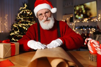 Photo of Santa Claus wrapping gift at his workplace in room decorated for Christmas