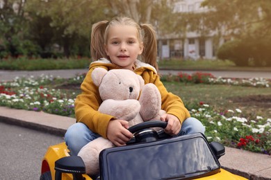 Cute little girl with toy bear driving children's car in park