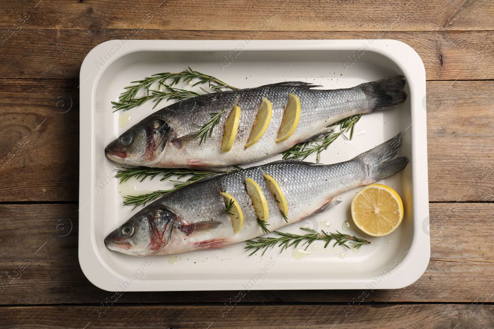Photo of Baking tray with raw sea bass fish, lemon and rosemary on wooden table, top view