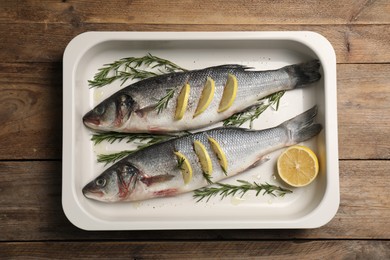 Photo of Baking tray with raw sea bass fish, lemon and rosemary on wooden table, top view