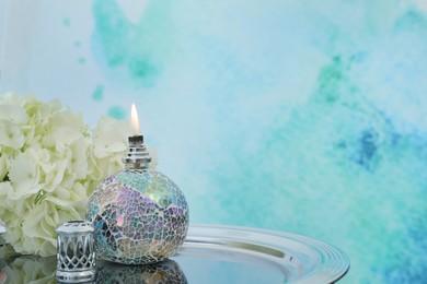 Photo of Burning catalytic lamp with hydrangea on tray against light blue background, space for text. Element of interior
