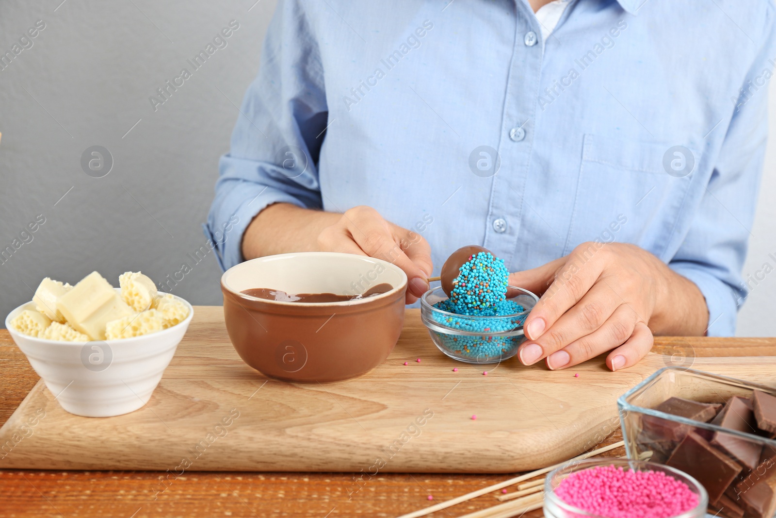 Photo of Young woman putting cake pop into blue sprinkles on wooden board, closeup