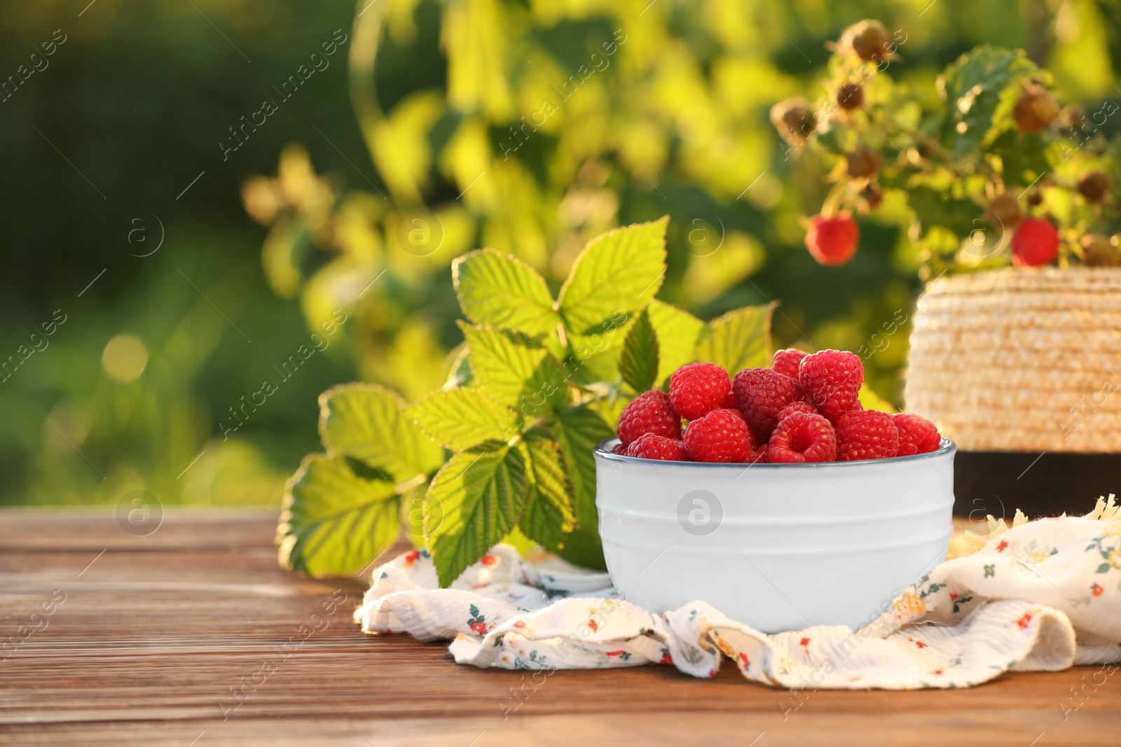 Photo of Tasty ripe raspberries in bowl, green leaves and straw hat on wooden table outdoors, space for text