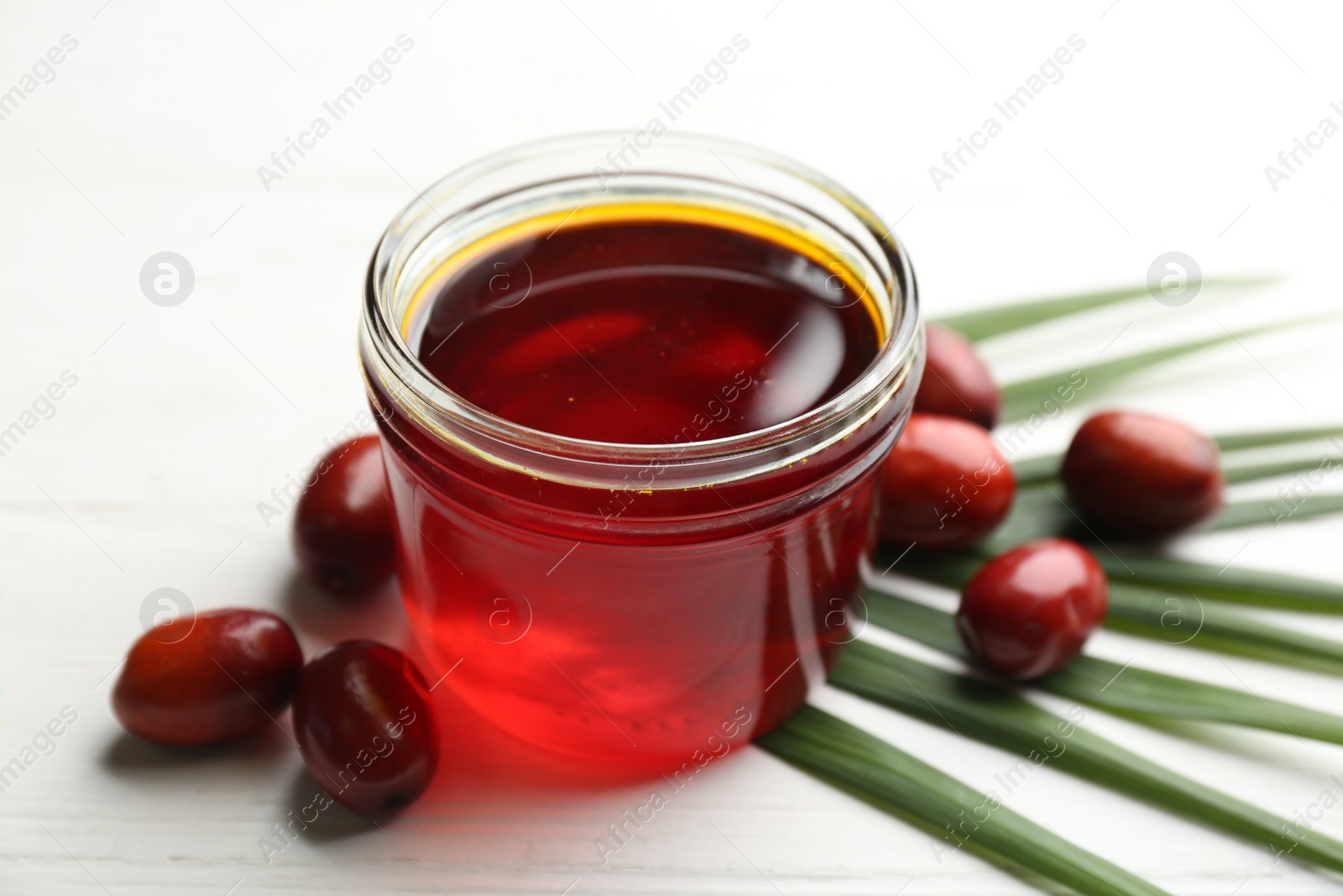 Photo of Palm oil in glass jar, tropical leaf and fruits on white wooden table