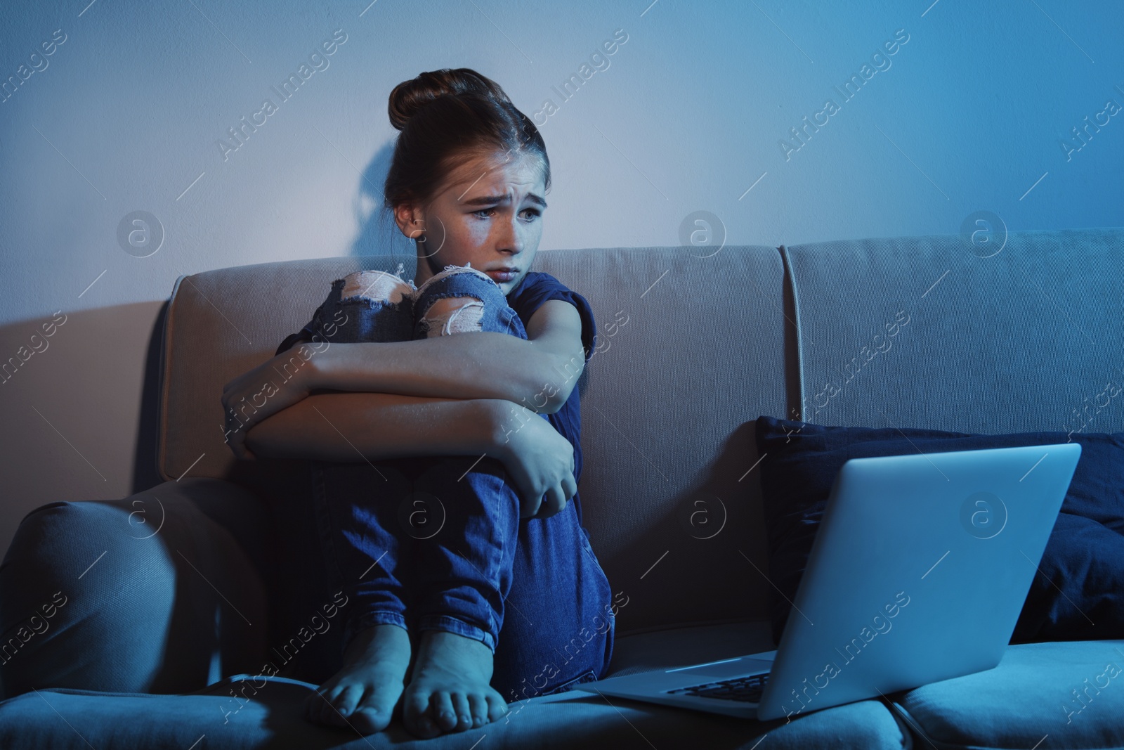 Photo of Frightened teenage girl with laptop on sofa in dark room. Danger of internet