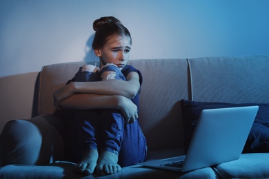 Photo of Frightened teenage girl with laptop on sofa in dark room. Danger of internet