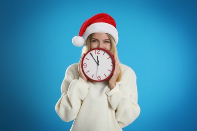 Woman in Santa hat with clock on light blue background. New Year countdown