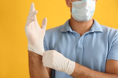 Man in protective face mask putting on medical gloves against yellow background, closeup