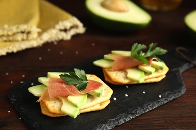 Photo of Delicious crackers with avocado, prosciutto and parsley on wooden table, closeup