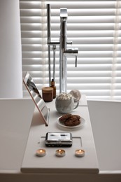 Photo of White wooden tray with tablet, spa products and burning candles on bathtub in bathroom