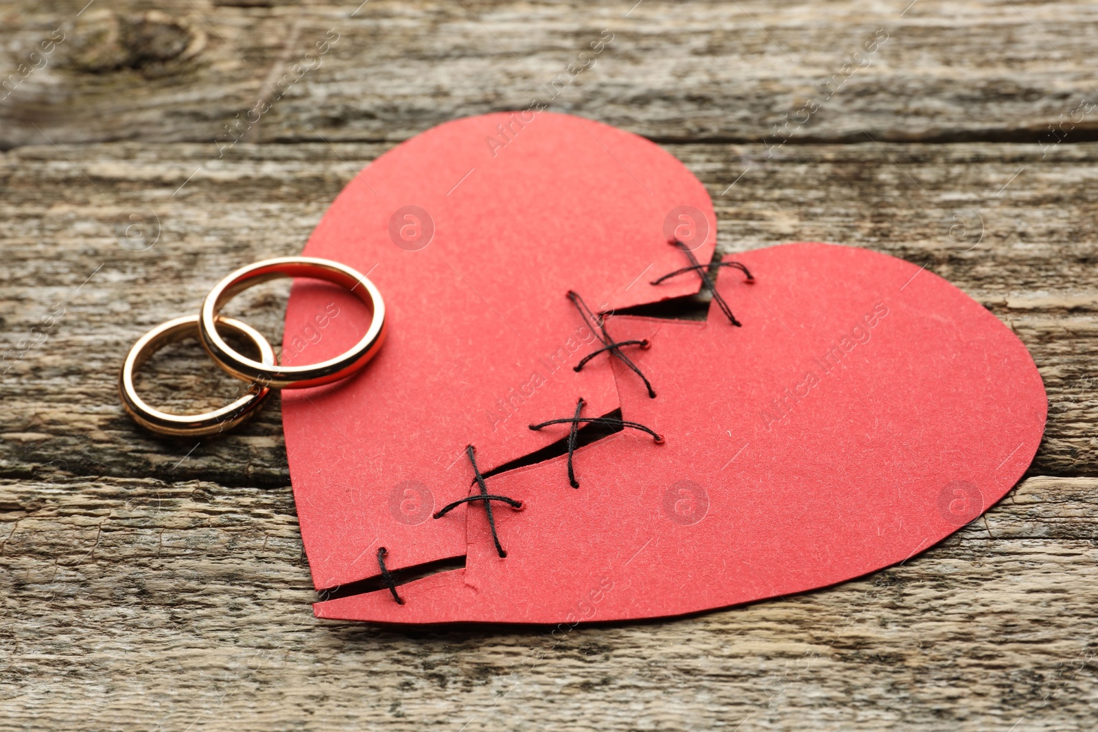 Photo of Broken heart. Torn red paper heart sewed with thread and wedding rings on wooden table, closeup